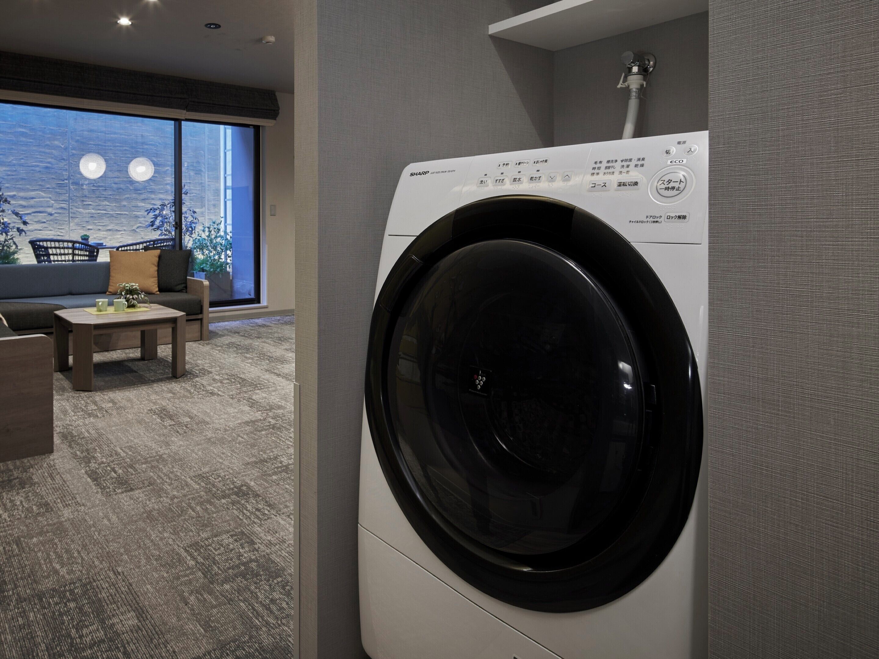 A modern laundry room with a front-loading washing machine, a sofa, a coffee table, and a large window with a view of the moon.