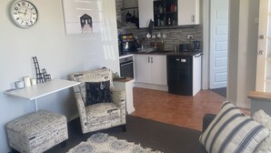 A compact living space with a kitchenette, a dining area with a white table and two chairs, a patterned armchair, and a decorative rug. - Self Contained Unit (Eaton)