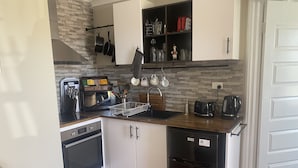 A compact kitchen with white cabinets, a black refrigerator, a built-in oven, a coffee maker, and a hanging pot rack. - Self Contained Unit (Eaton)