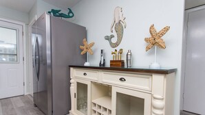 Una cocina con un refrigerador de acero inoxidable, un mueble color crema con cajones y estantes, y decoración de paredes con estrellas de mar y figuras de sirenas.