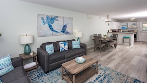 Un salón con un sofá gris, una mesa de centro de madera y una alfombra azul y blanca estampada. Hay una zona de cocina con una mesa y sillas de comedor.