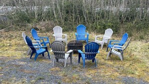 A group of Adirondack chairs arranged around a small round table in an outdoor setting with a forest background. - Spacious Home on a Rushing Glacier Fed Creek (Seward)