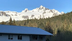 A two-story house with a sloped roof, a front porch, and a garage. It is surrounded by a forest and has a mountain range in the background. - Spacious Home on a Rushing Glacier Fed Creek (Seward)