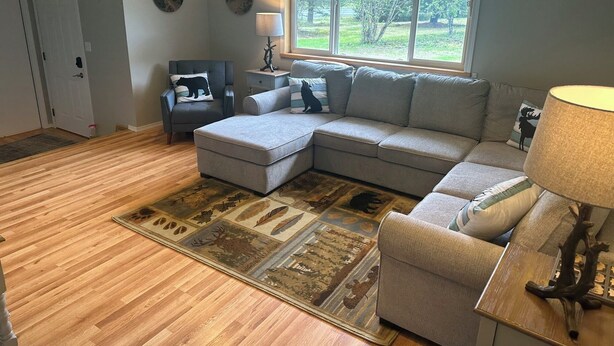 A living room with a sectional sofa, a small chair, a rug, a lamp, and a window with a view of trees. - Spacious Home on a Rushing Glacier Fed Creek (Seward)