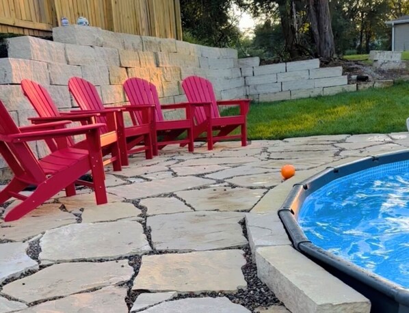 Un jardin avec une terrasse en pierre, une piscine et des fauteuils Adirondack rouges.