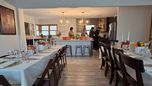 A dining area with a long table set for a meal, a kitchen in the background, and a bar area with people.