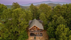 Une cabane en rondins à deux étages, dotée de fondations en pierre, entourée d’une forêt dense, et comprenant un balcon à l’étage supérieur et une véranda à l’avant.