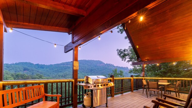 Une terrasse en bois avec un barbecue et un espace salon, offrant une vue sur un paysage boisé.