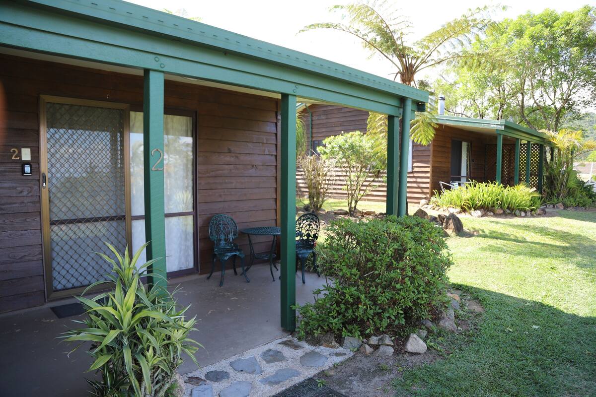 A wooden building with a covered porch, two chairs, and a small garden area with plants and rocks.
