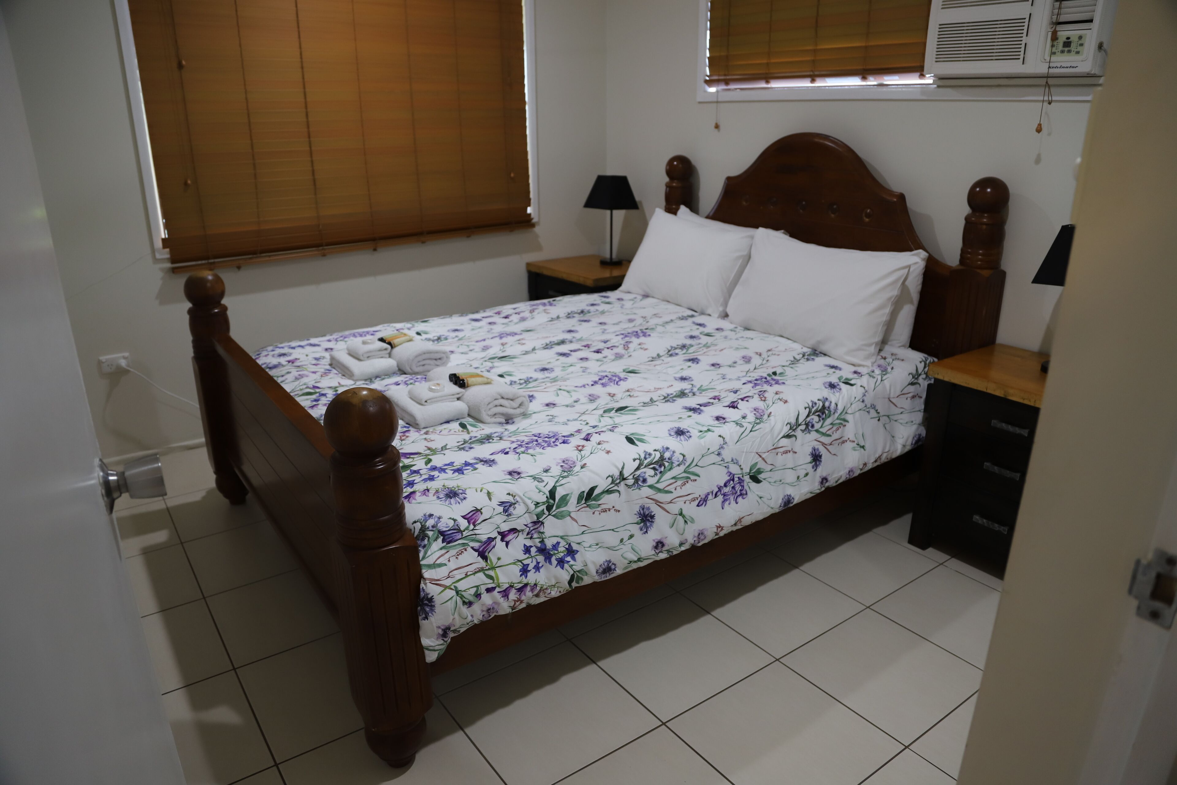 A neatly made bed with a floral bedspread, two bedside tables with lamps, a wooden headboard, and a window with blinds.