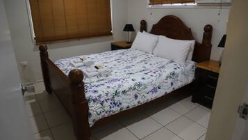 A neatly made bed with a floral bedspread, two bedside tables with lamps, a wooden headboard, and a window with blinds.