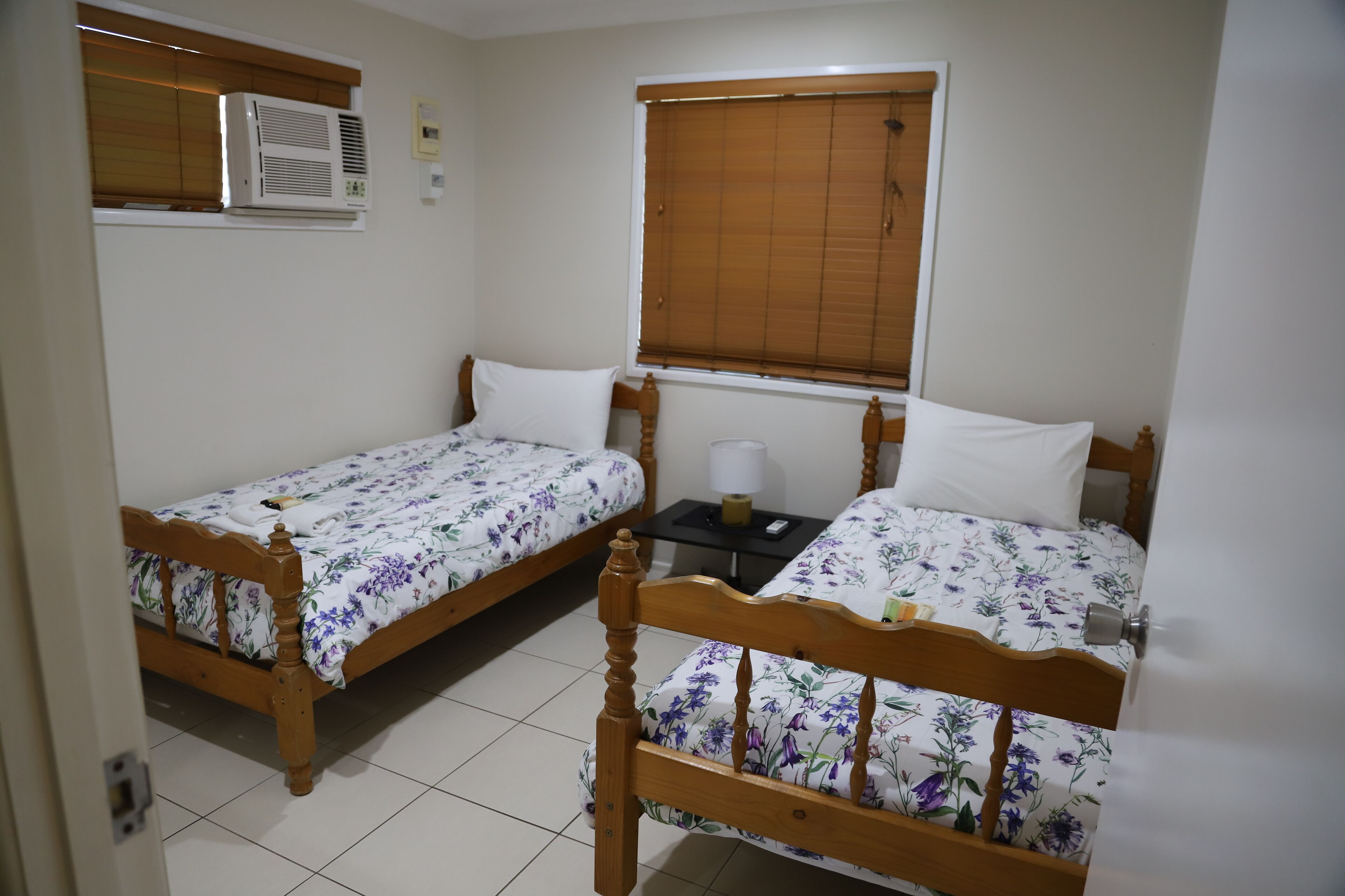A bedroom with two wooden beds, a window with blinds, an air conditioning unit, and a small bedside table with a lamp.