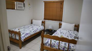 A bedroom with two wooden beds, a window with blinds, an air conditioning unit, and a small bedside table with a lamp.