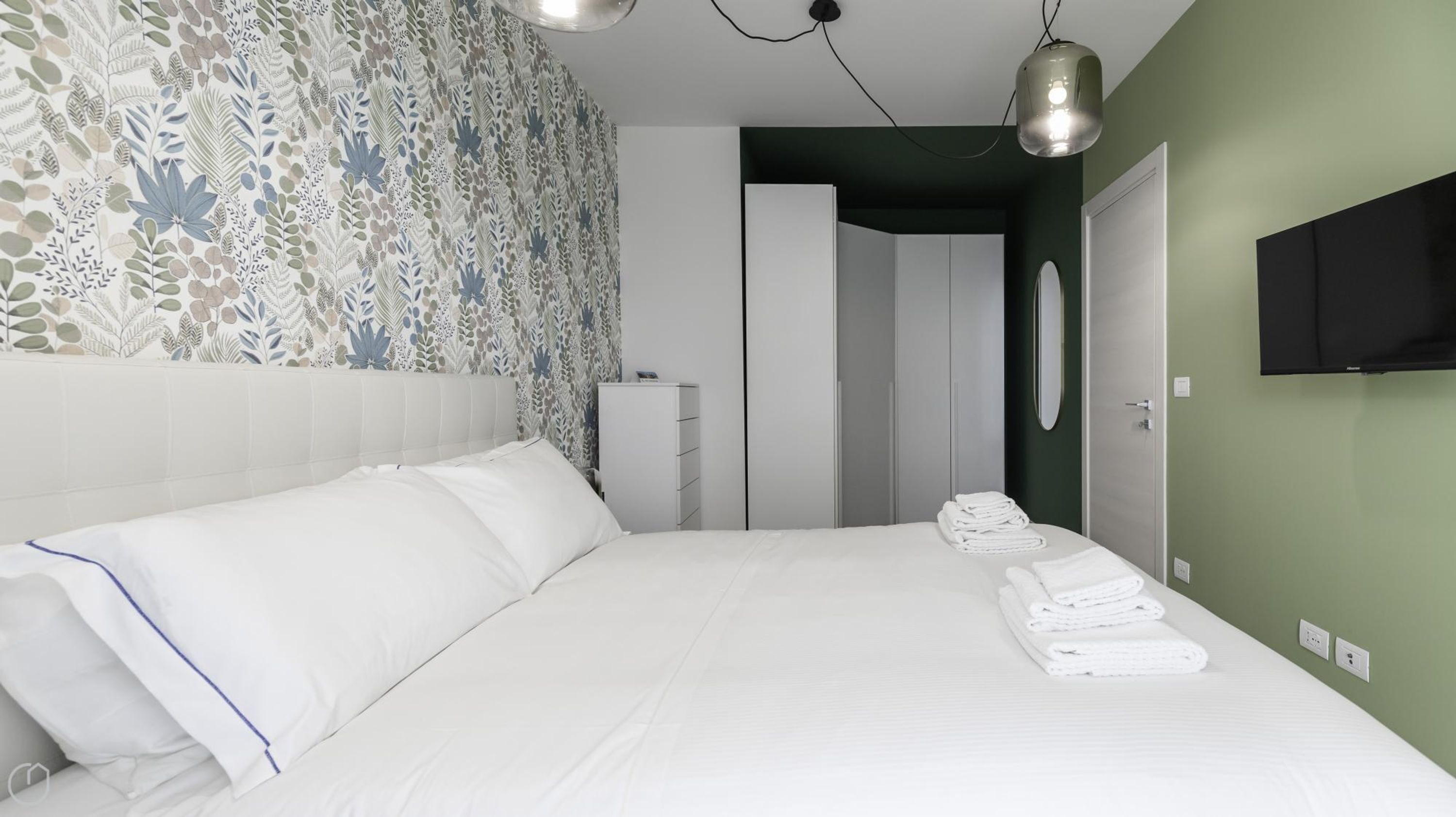 A neatly made bed with white linens and a blue accent, a white dresser, a wardrobe, and a flat-screen TV mounted on the wall.