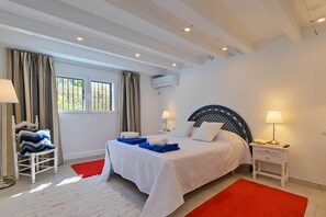 A bedroom with a bed, bedside tables, a chair, a window with curtains, and an air conditioning unit.