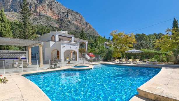 Una piscina amb muntanyes al fons, un edifici blanc amb finestres d'arc i mobiliari exterior.