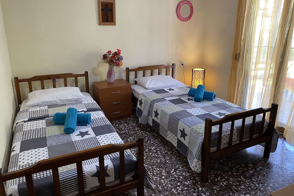 A bedroom with two beds, a bedside table with a vase of flowers, a wall-mounted cross, and a window with curtains.