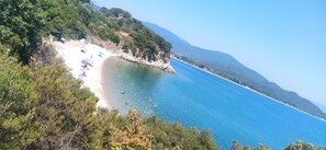 A coastal beach with clear blue water, surrounded by greenery and hills.
