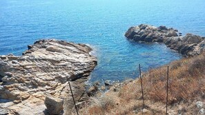 A rocky coastline with clear blue sea, a fenced area, and sparse vegetation.