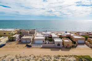 A coastal residential area with multiple buildings, a sandy foreground, and a clear view of the sea.