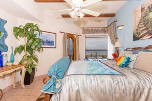 A bedroom with a bed, a ceiling fan, a window with curtains, a plant, and a painting on the wall.