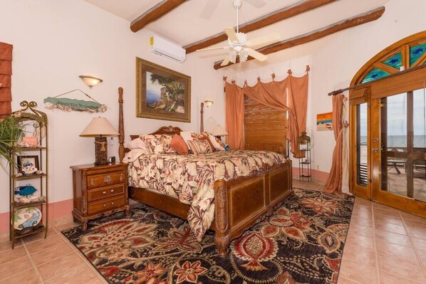 A bedroom with a bed, a nightstand, a dresser, a ceiling fan, a window with curtains, and a view of the sea.