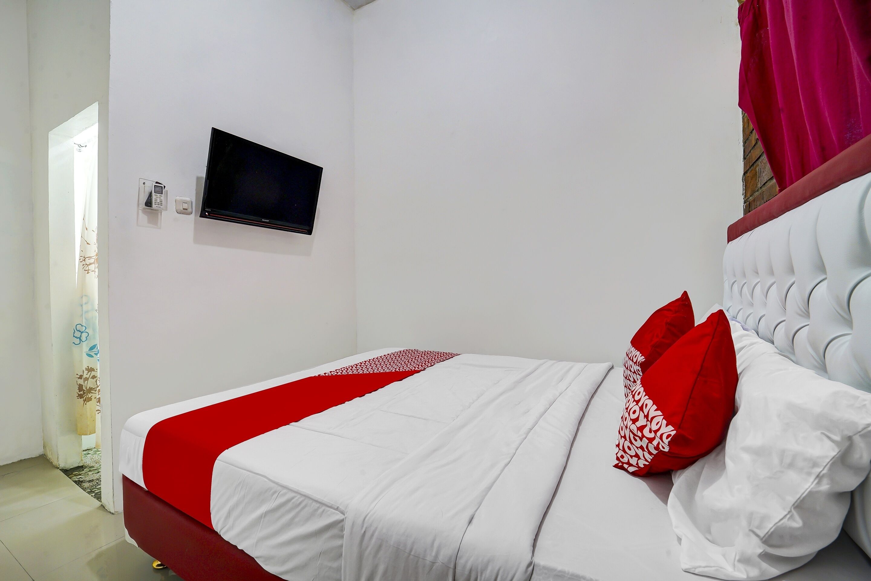 A neatly made bed with white and red bedding, a flat-screen TV mounted on the wall, and a red curtain on the window.