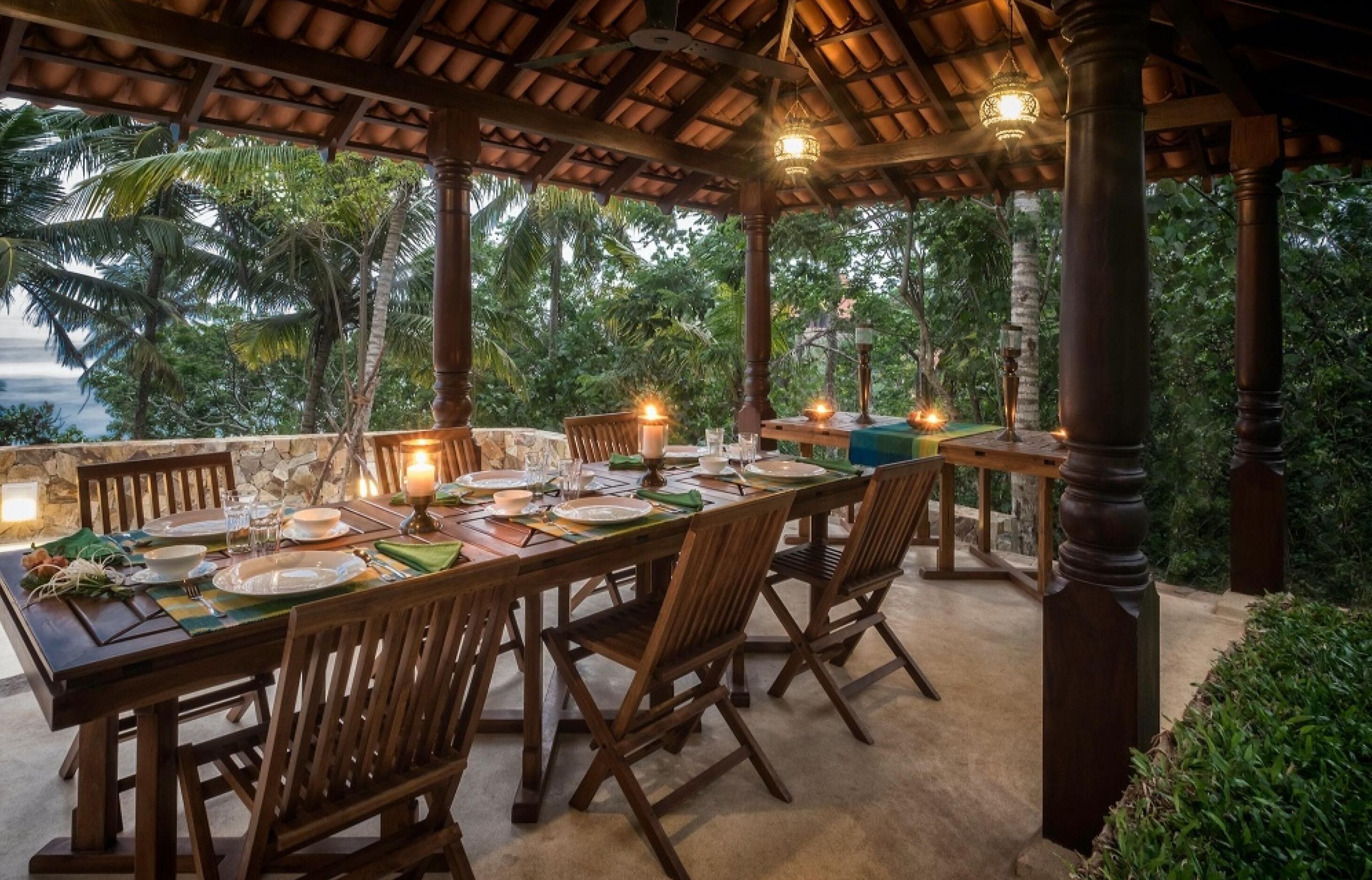 A covered dining area with a long table set for a meal, surrounded by wooden chairs, and featuring a view of palm trees and a body of water.