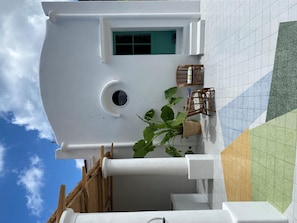 Un edificio blanco con un ventanal redondo, una puerta con marco verde y un pequeño balcón con mobiliario de mimbre y una planta.