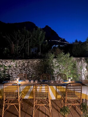 An outdoor dining area with wicker chairs and a table set for four, surrounded by a stone wall and greenery, overlooking a mountainous landscape at night. - Capri Designer Suite by Elite Villas (Capri)