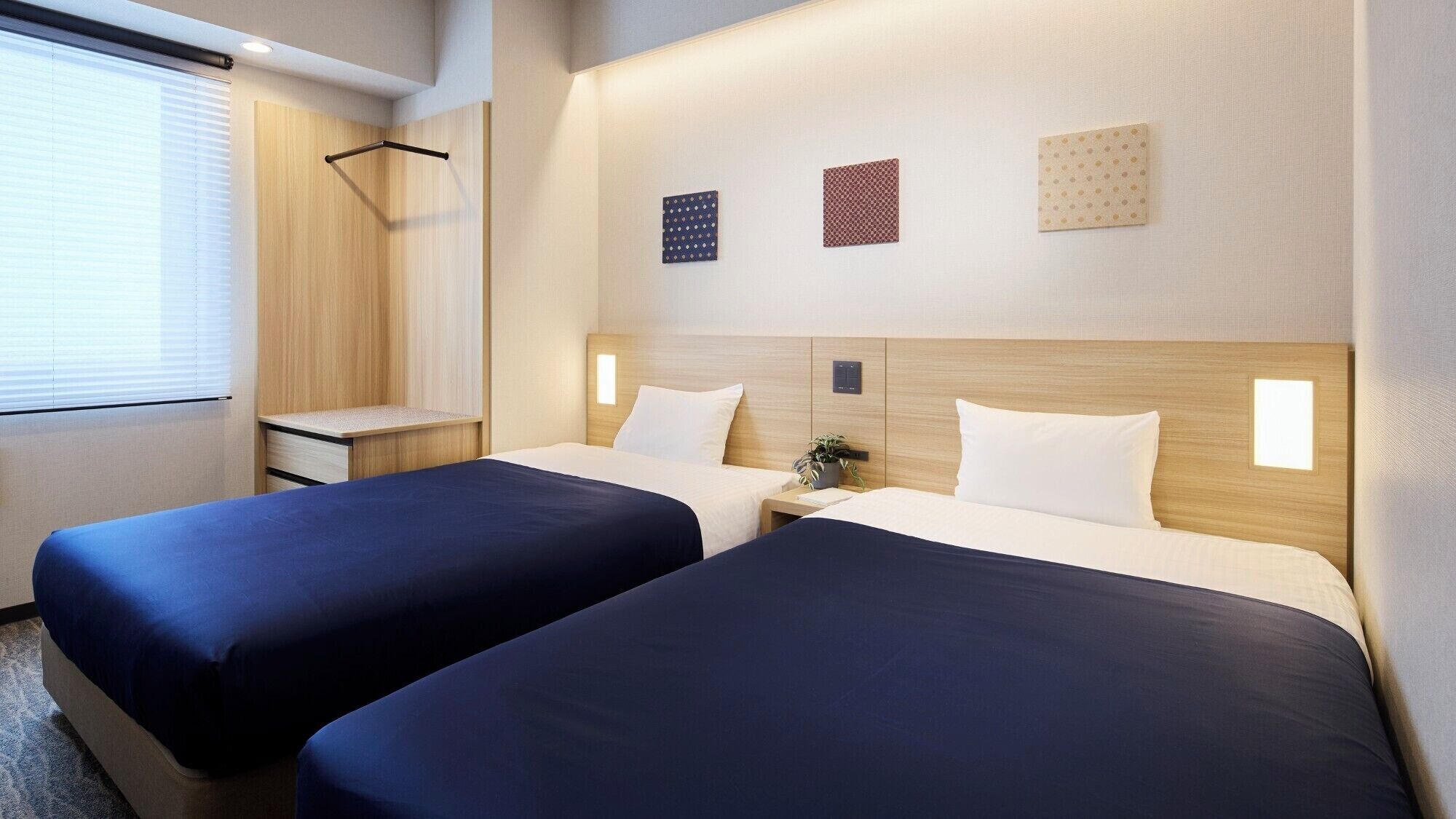 A hotel room with two beds, a small bedside table, a wall-mounted light, and a window with blinds.