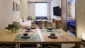 A dining area with a wooden table set for four, a sofa, a TV, and a window with blinds.