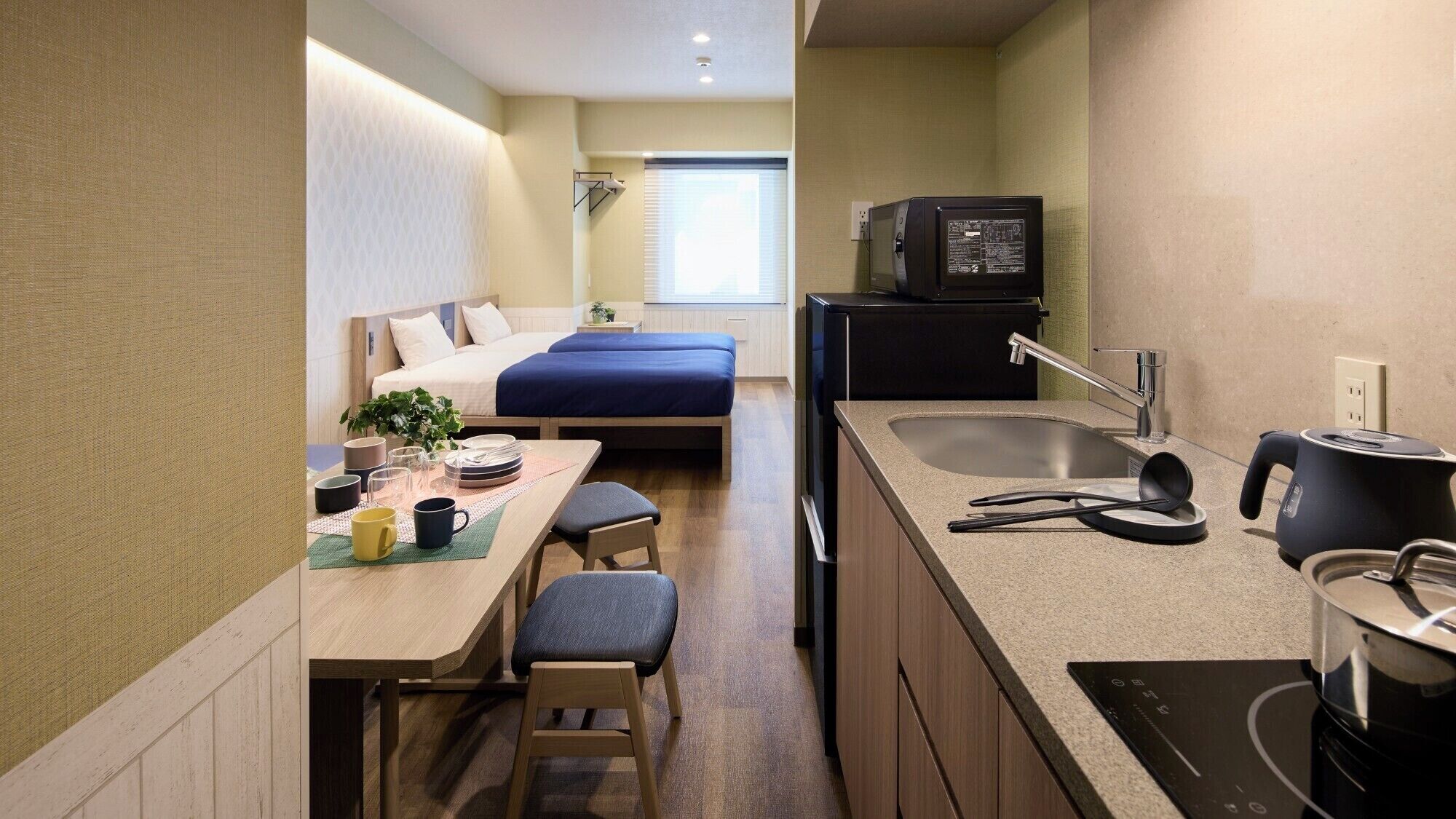 Een compacte hotelkamer met een kitchenette, een bed met blauw beddengoed, een eettafel met stoelen en een klein raam met zonwering.