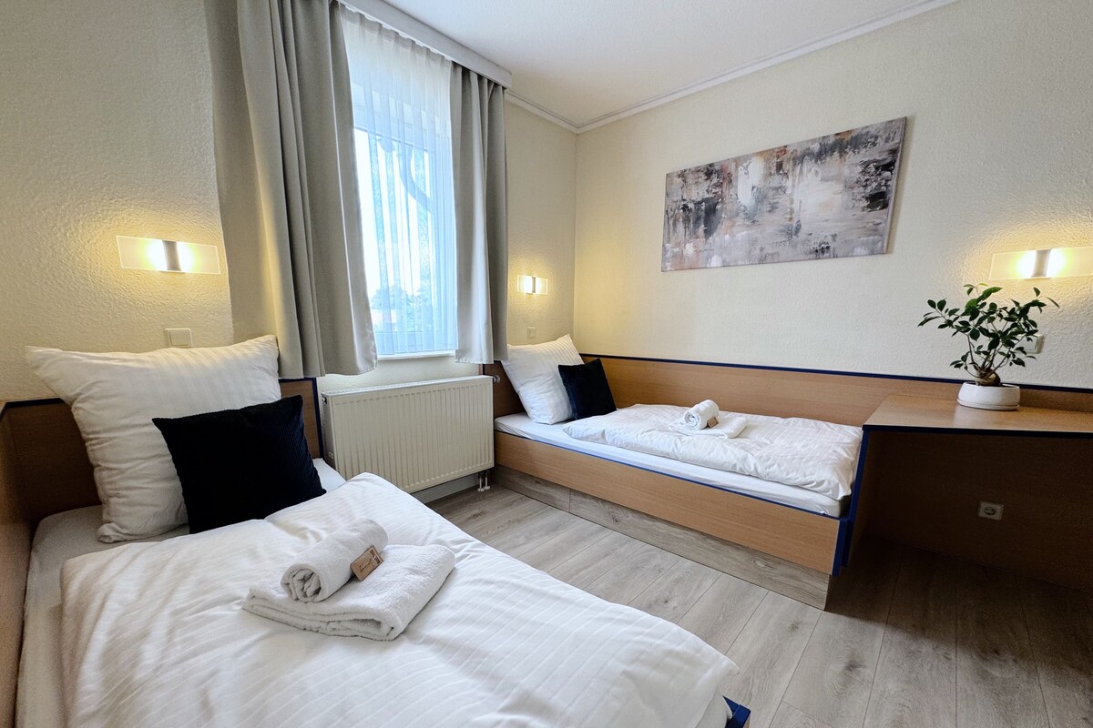A hotel room with two beds, a window with curtains, a radiator, a painting on the wall, and a potted plant.