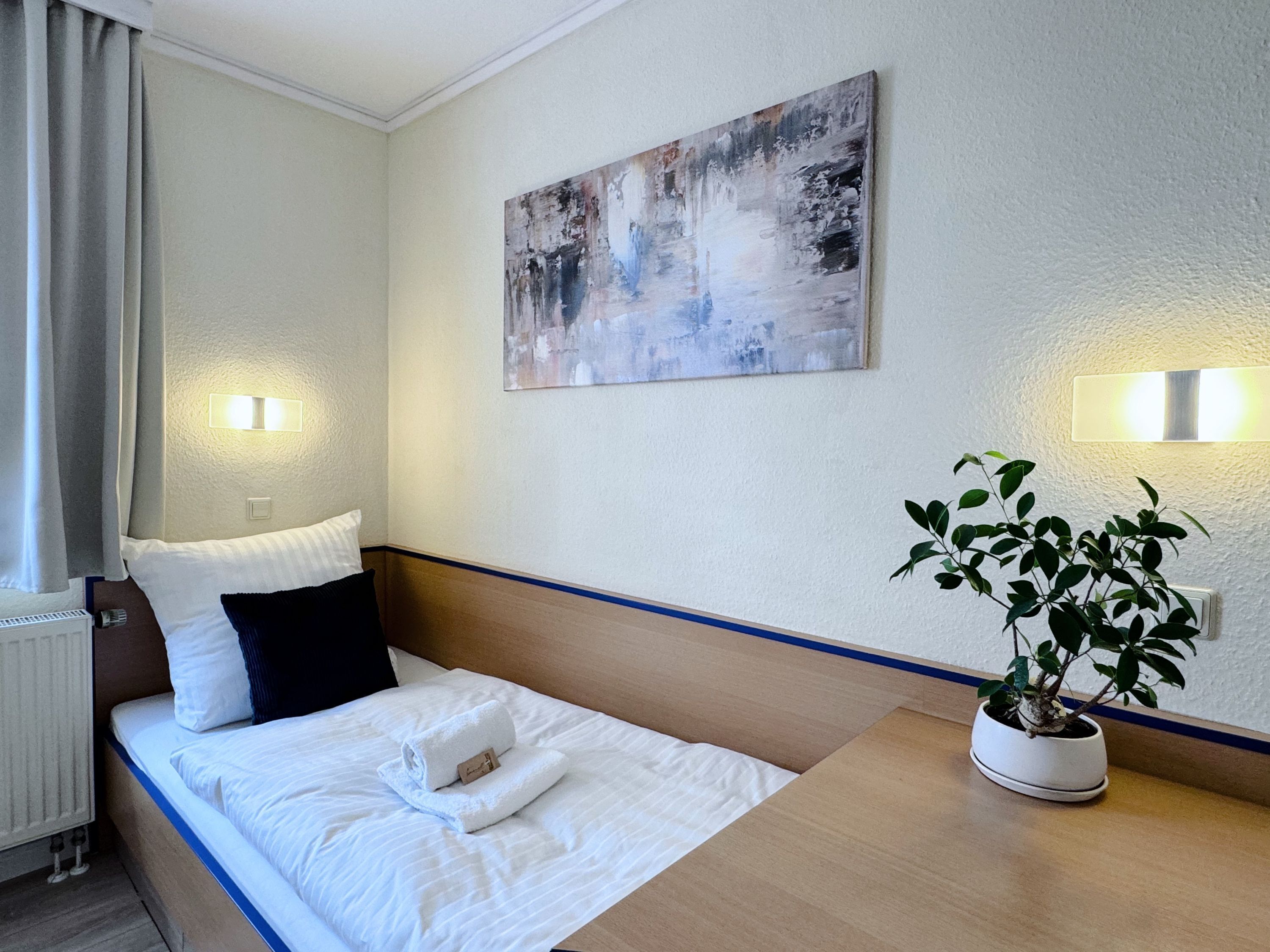Una habitación de hotel con una cama, una mesita de noche con una planta en maceta, un cuadro en la pared y dos luces fijadas a la pared.