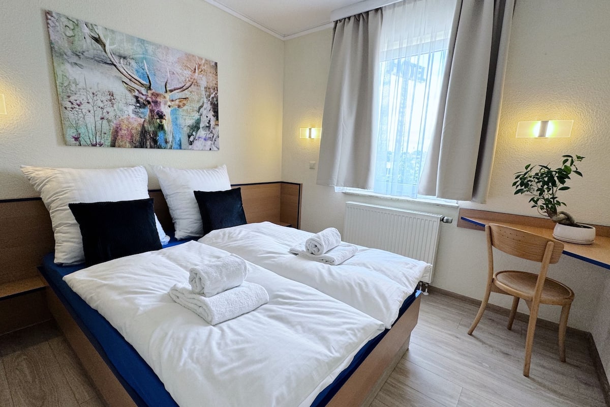 A hotel room with a large bed, a desk, a chair, a window with curtains, a radiator, and a painting on the wall.