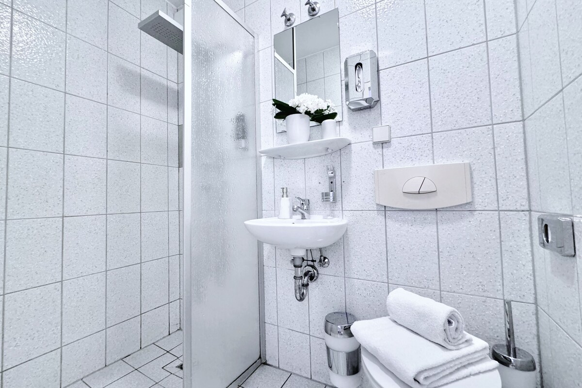 A modern bathroom with a glass shower enclosure, a white sink with a mirror above it, and a toilet with a towel stack beside it.