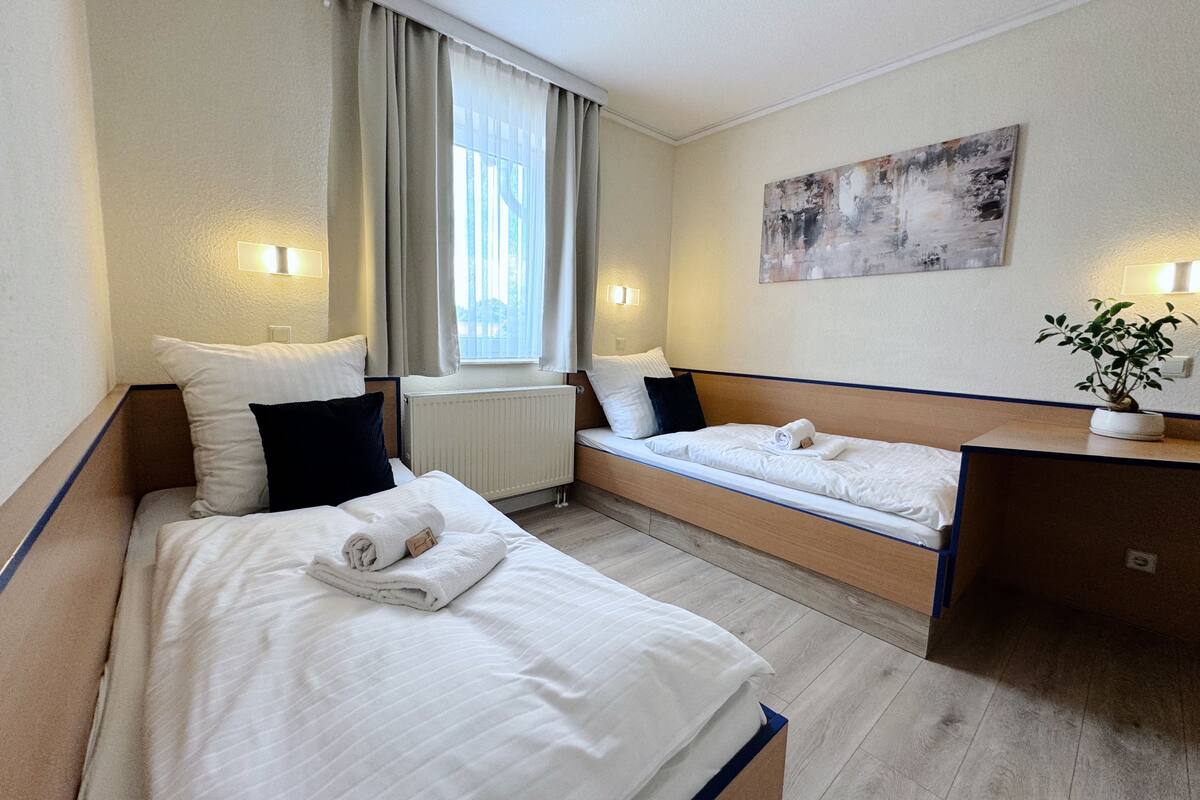 A hotel room with two beds, a desk, a radiator, and a painting on the wall.