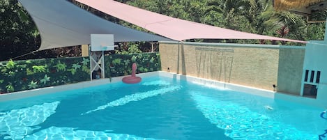 An outdoor swimming pool with clear water, surrounded by lush greenery and a shaded area.