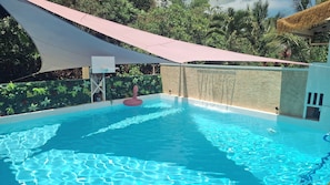 Una piscina exterior con agua cristalina, rodeada de abundante vegetación y una zona con sombra.