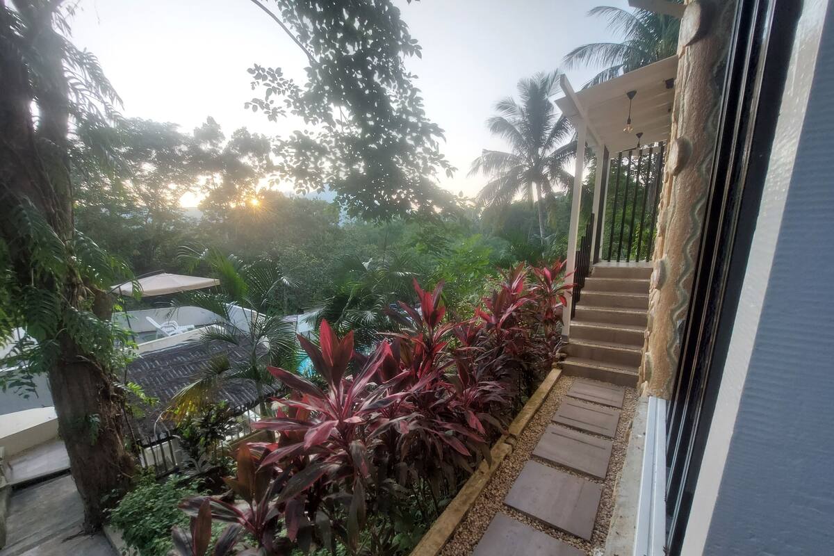 A balcony with a view of a garden, a pool, and a staircase leading to an upper level.