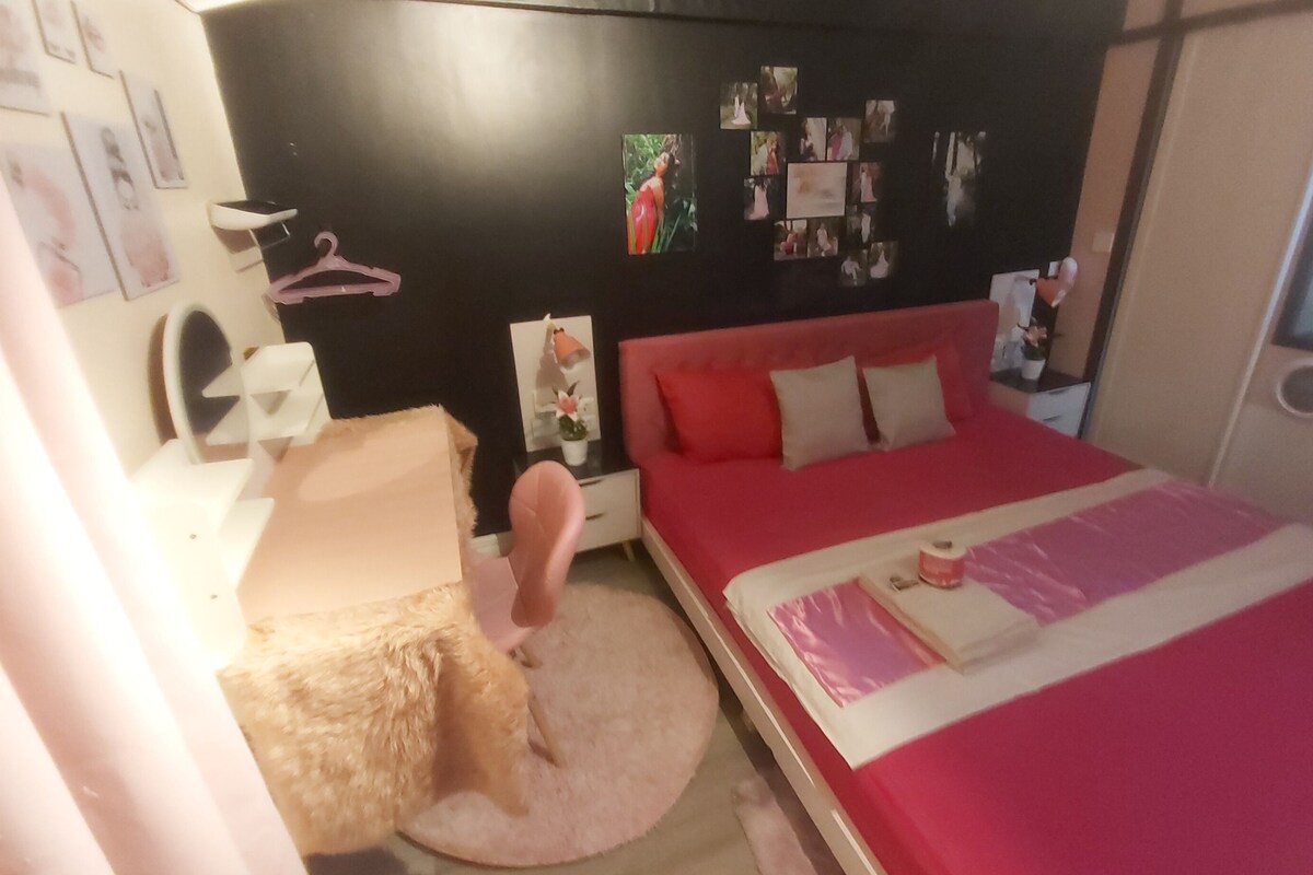 Un dormitorio con una cama roja, una mesita de noche blanca, una silla rosa y una pared con fotografías.