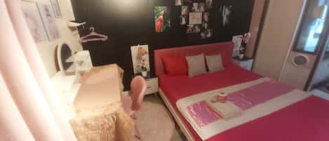 Un dormitorio con una cama roja, una mesita de noche blanca, una silla rosa y una pared con fotografías.