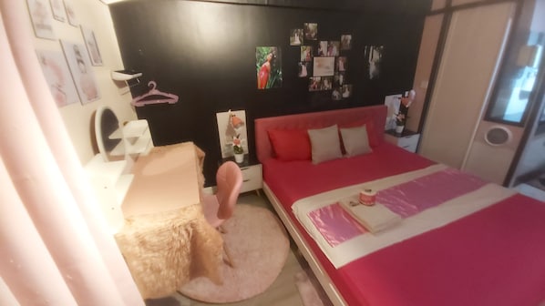 Une chambre avec un lit rouge, une table de chevet blanche, une chaise rose et un mur décoré de photos.