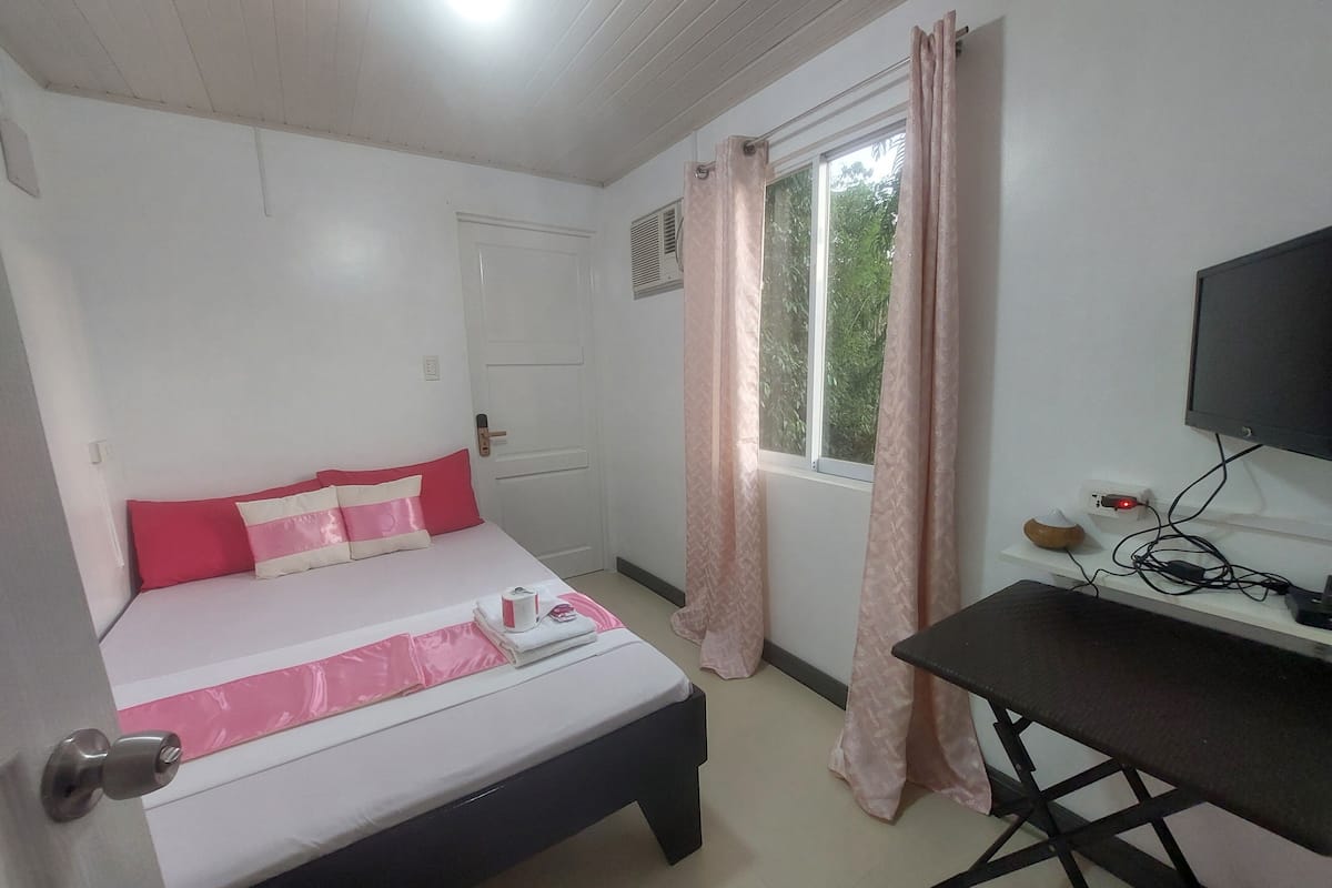 A bedroom with a bed, a desk, a TV, and a window with curtains.
