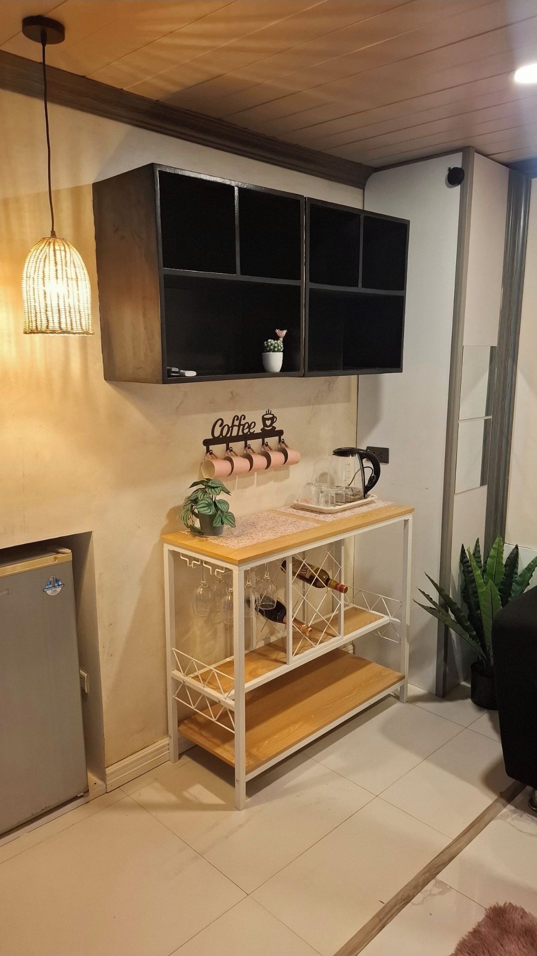 A room with a wooden ceiling, a wall-mounted shelf with a coffee sign, a small table with a plant, wine glasses, and a bottle.