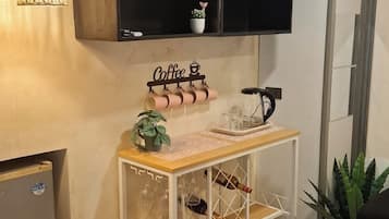 A room with a wooden ceiling, a wall-mounted shelf with a coffee sign, a small table with a plant, wine glasses, and a bottle.