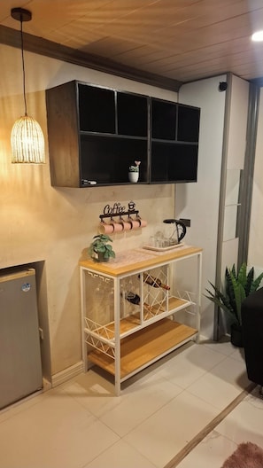 Une pièce avec un plafond en bois, une étagère fixée au mur sur laquelle est posé un panneau indiquant « café », une petite table avec une plante, des verres à vin et une bouteille.