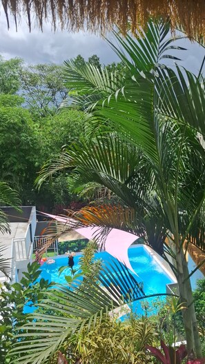 Une piscine entourée d’une végétation luxuriante, une structure blanche dotée d’un toit et un ciel bleu azur.