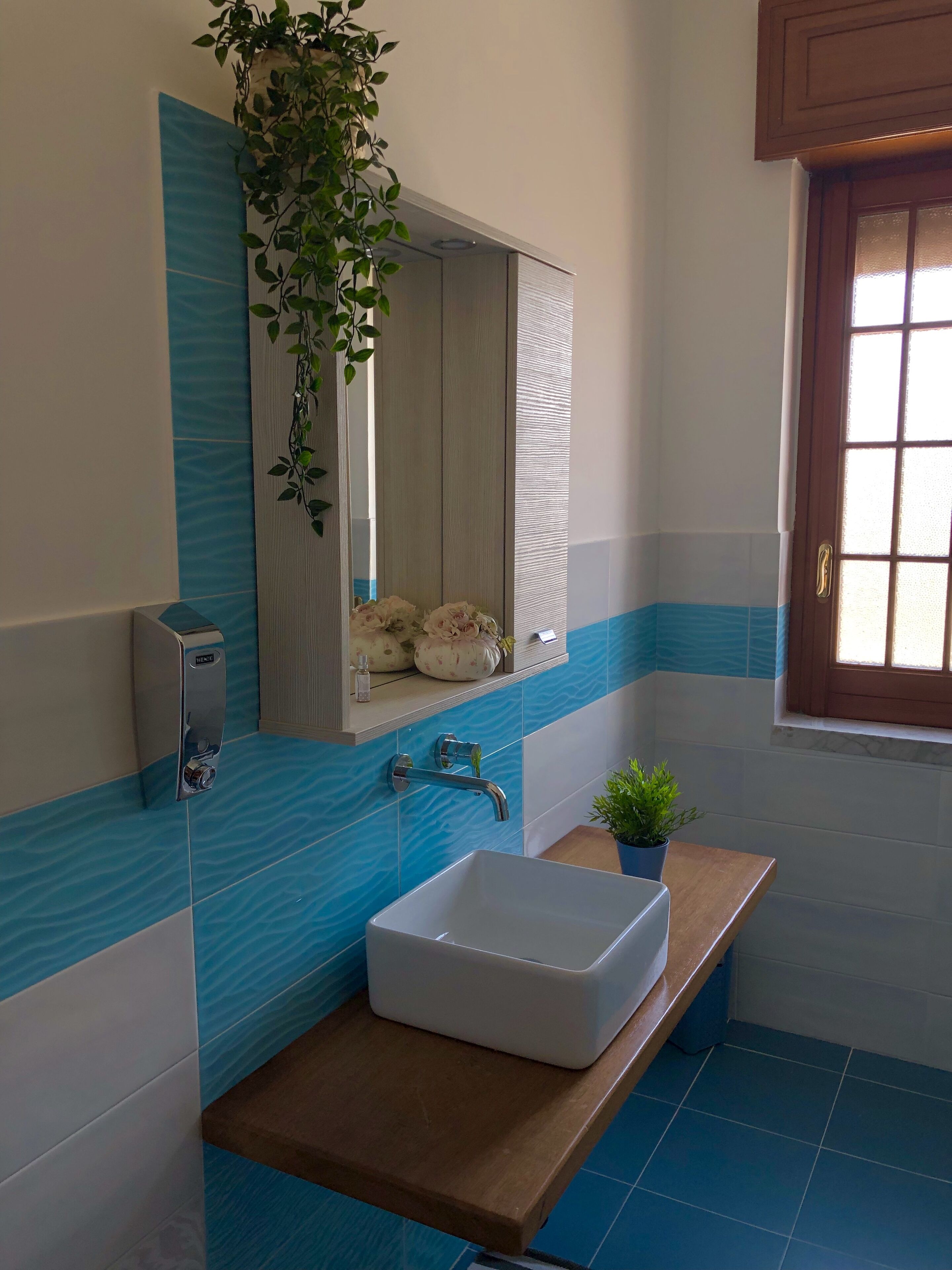 A bathroom with a white sink, blue-tiled walls, a wooden countertop, a mirror, a hanging plant, and a window with blinds.
